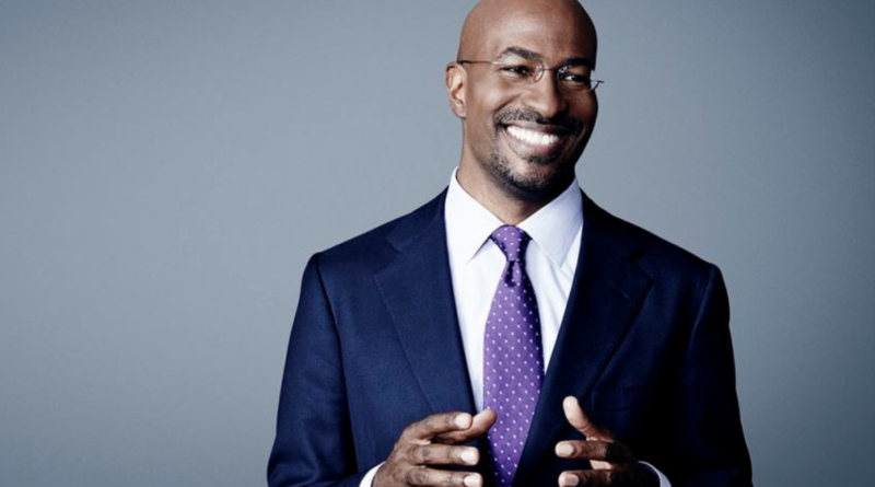 A man in a suit and tie smiles while gesturing with his hands against a neutral studio background.
