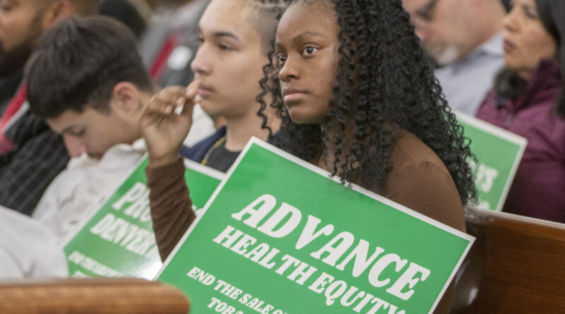 African American teenager sits with peers in meeting, holds "Advance Health Equity" sign with white text over green background.
