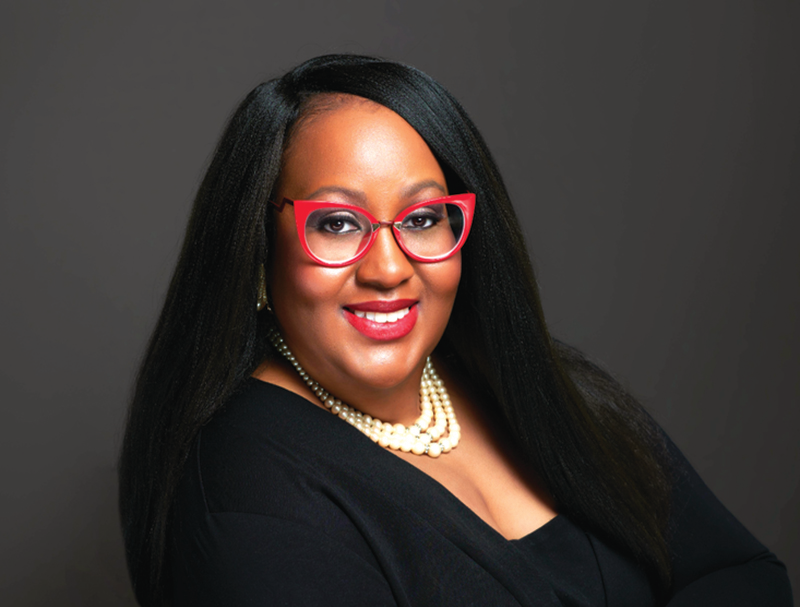 A Black woman with long Black hair wearing red framed glasses, pearls, and a black top.