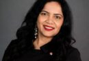 Woman with dark hair and red lipstick smiles at camera with gray background.