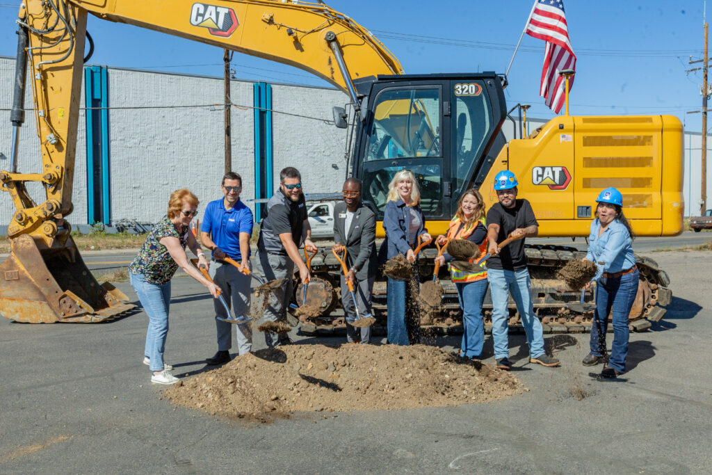 Group of people standing in front of an excavator and behind a small mound of dirt holding shovels to signify the groundbreaking of a construction project.