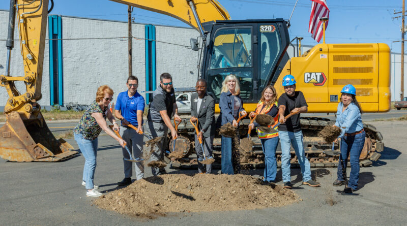 Group of people standing in front of an excavator and behind a small mound of dirt holding shovels to signify the groundbreaking of a construction project.