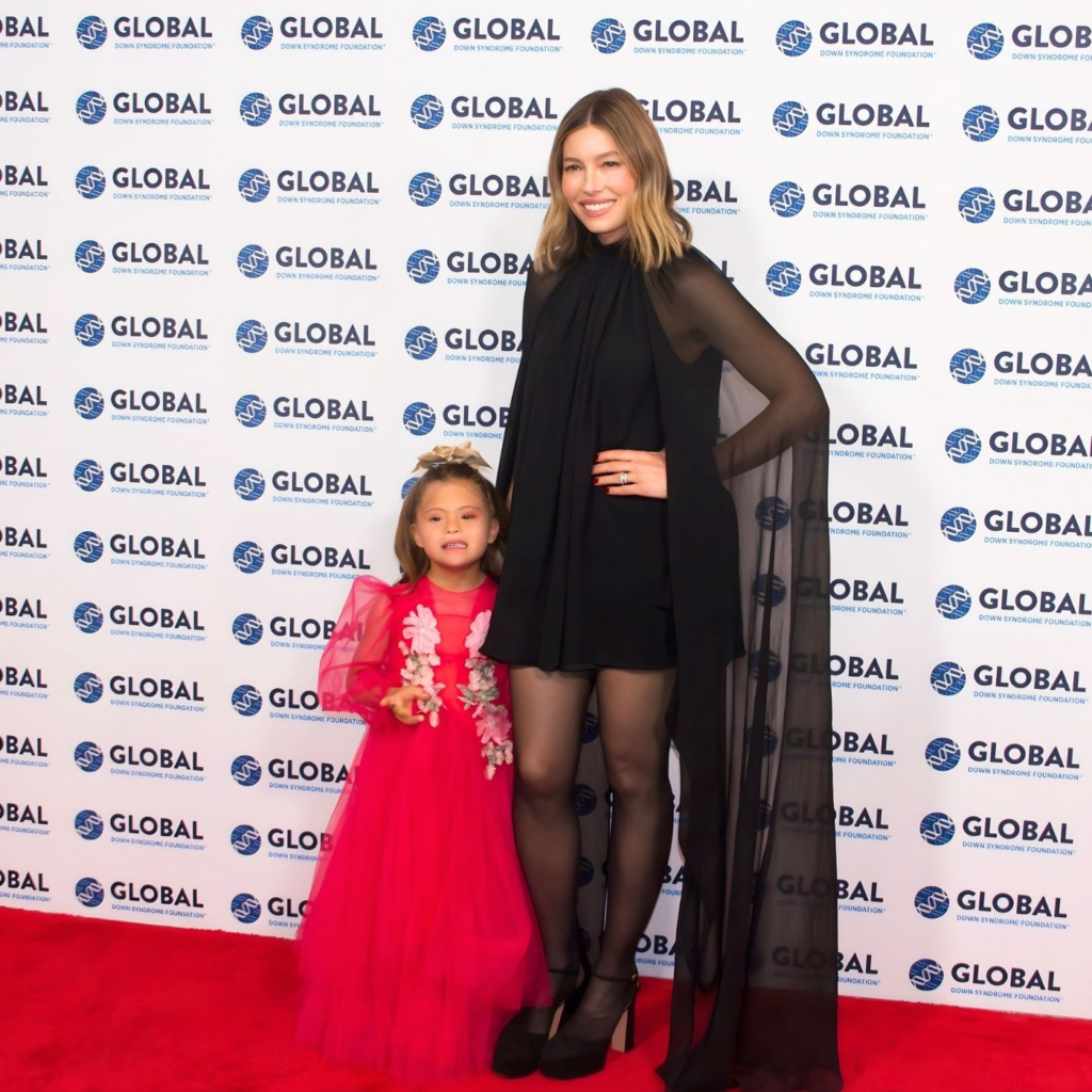 Young girl (Zaya Biel) wearing bright pink dress, standing next to Jessica Biel who is wearing a black dress with sheer cape. 