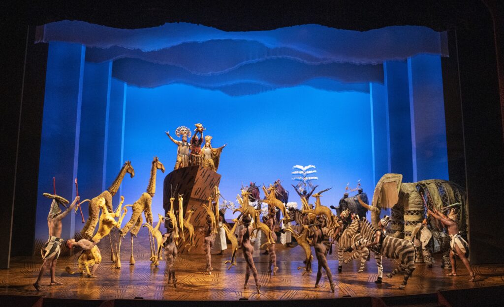 A vibrant stage scene from The Lion King musical, featuring actors in elaborate animal costumes—giraffes, zebras, elephants, and birds—gathered around Pride Rock as Rafiki raises baby Simba under a glowing blue sky backdrop.