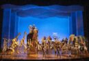 A vibrant stage scene from The Lion King musical, featuring actors in elaborate animal costumes—giraffes, zebras, elephants, and birds—gathered around Pride Rock as Rafiki raises baby Simba under a glowing blue sky backdrop.