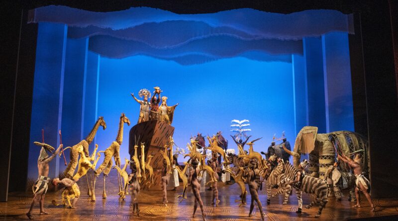 A vibrant stage scene from The Lion King musical, featuring actors in elaborate animal costumes—giraffes, zebras, elephants, and birds—gathered around Pride Rock as Rafiki raises baby Simba under a glowing blue sky backdrop.