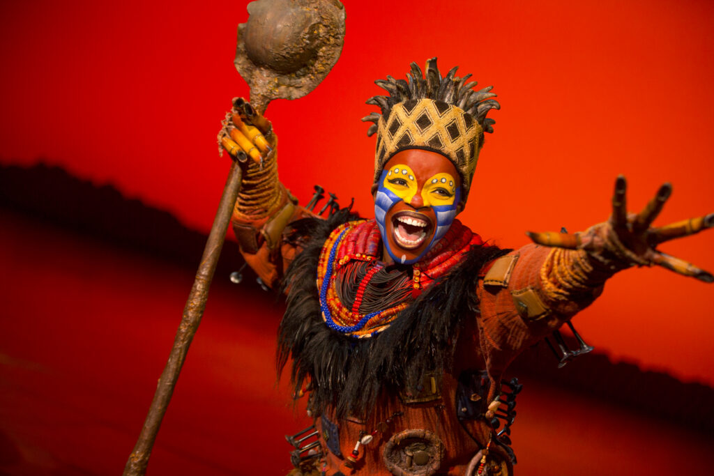 A performer portraying Rafiki in The Lion King musical smiles joyfully while holding a staff, wearing vibrant face paint, colorful beads, and an ornate headdress against a bold red stage backdrop.