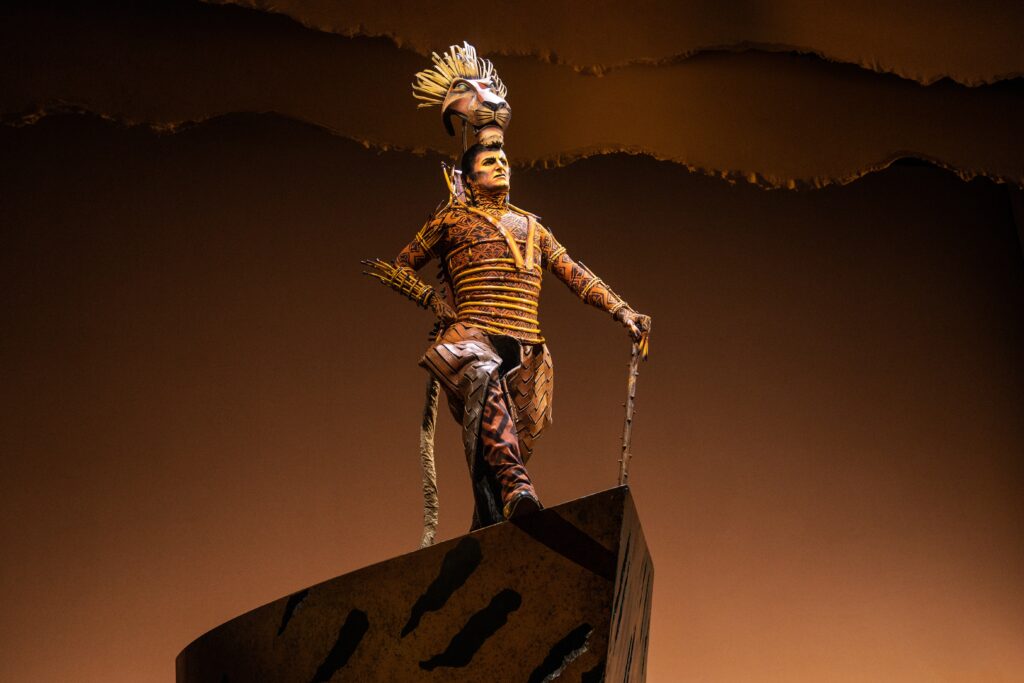 An actor portraying Scar in The Lion King musical stands atop a rock-like platform, wearing an intricate costume of earth tones and a lion-inspired headpiece, lit against a warm amber backdrop.