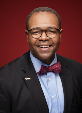 A smiling man wearing glasses, a dark suit jacket, light blue shirt, and red bow tie poses in front of a dark red background.