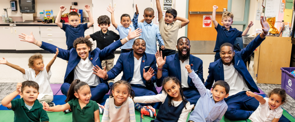 A group of elementary school children and four adult mentors wearing blue suits sit together on a colorful classroom rug, smiling and raising their hands in celebration. The joyful scene captures a moment of connection and encouragement between students and mentors in an educational setting.