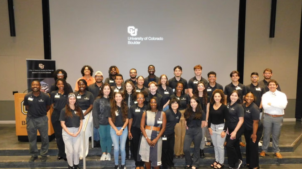 A large group of diverse students and faculty pose together on the steps of an auditorium at the University of Colorado Boulder, smiling in front of a projection screen displaying the university’s logo.