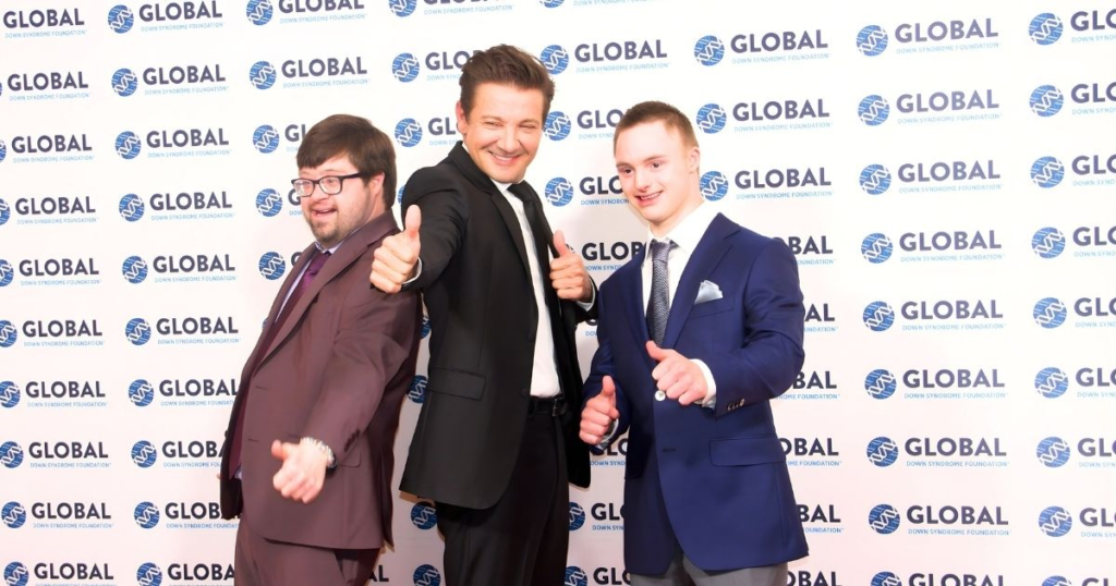 Global red carpet photo with Kevin Iannucci (L) a man with brown hair wearing brown suit and dark rimmed glasses; Jeremy Renner (C - wearing black suit and tie with brown hair) and Guion Macsovits (R - blue suit). All three men holding thumbs up on GLOBAL red carpet