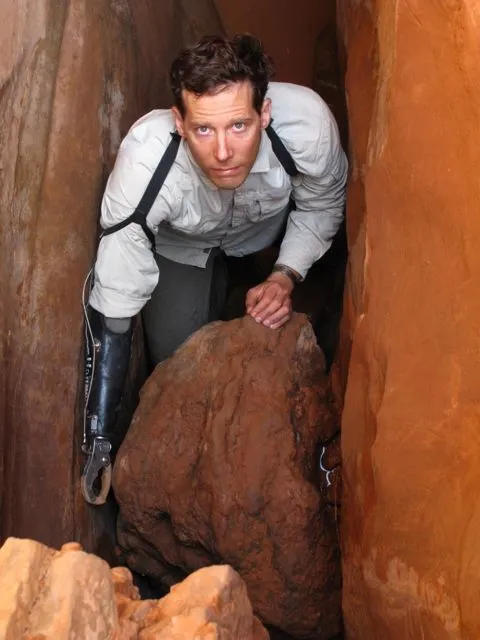 Aron Ralston stands inside a narrow red rock canyon, leaning forward and looking directly at the camera. His prosthetic right arm is visible as he braces himself against the canyon walls and a large boulder beneath him, referencing the location and circumstances of his well-known survival story.