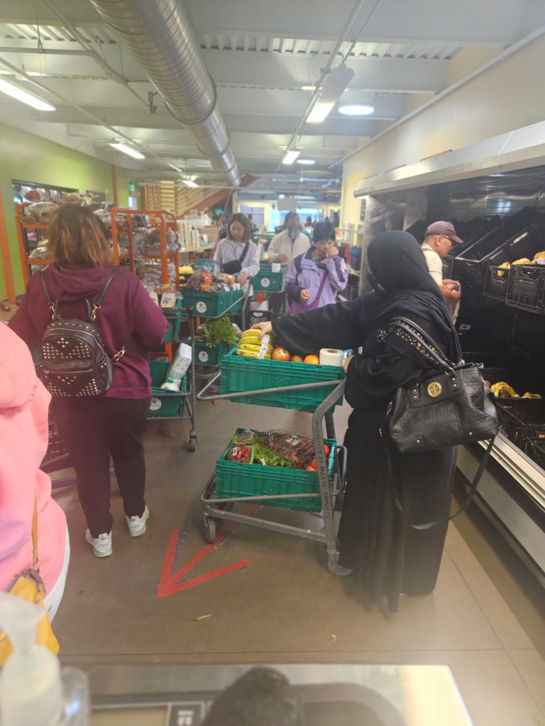 A crowded indoor food market space where several people stand in line with green plastic carts filled with fresh produce. Shoppers browse shelves and coolers along the walls, while overhead ductwork and fluorescent lighting highlight the busy atmosphere. A red arrow on the floor directs foot traffic. The scene reflects a high-demand day at a community food distribution center.