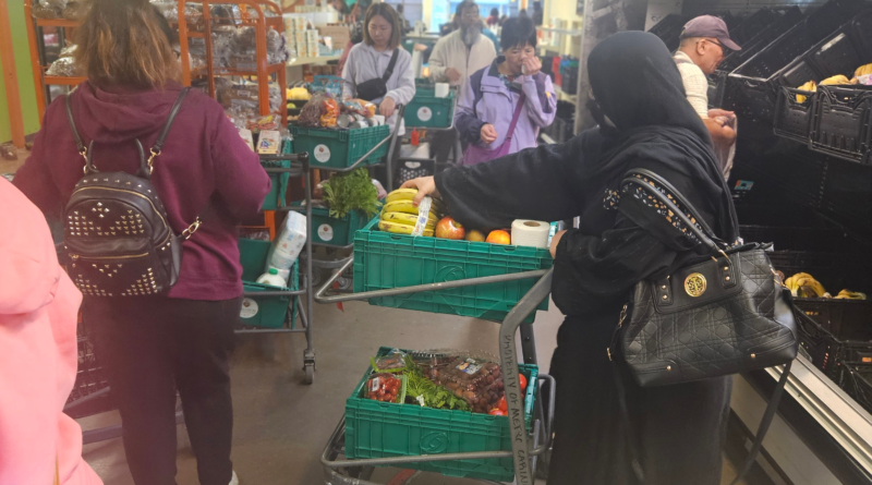 A crowded indoor food market space where several people stand in line with green plastic carts filled with fresh produce. Shoppers browse shelves and coolers along the walls, while overhead ductwork and fluorescent lighting highlight the busy atmosphere. A red arrow on the floor directs foot traffic. The scene reflects a high-demand day at a community food distribution center.