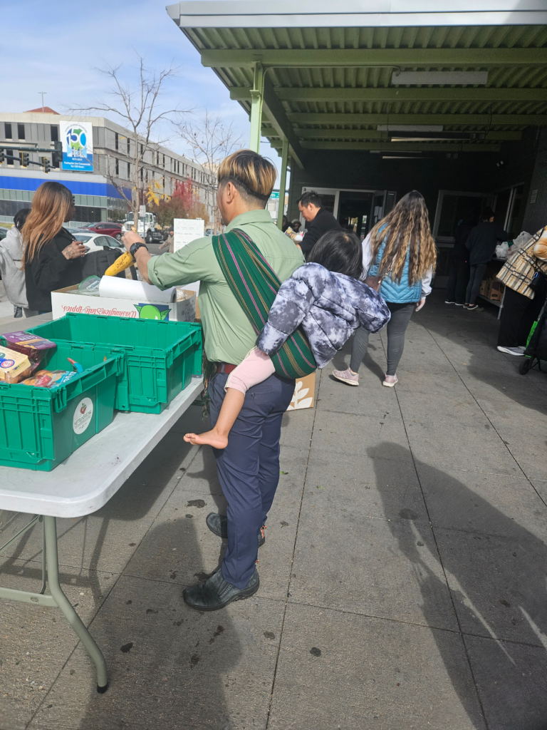 A parent stands outdoors at a food distribution table, selecting groceries from green plastic bins while carrying a small child on their back in a colorful sling. Other people gather around the tables in line, and a building with a green awning frames the scene. The atmosphere suggests a busy community food assistance event, with families receiving fresh food and pantry items.