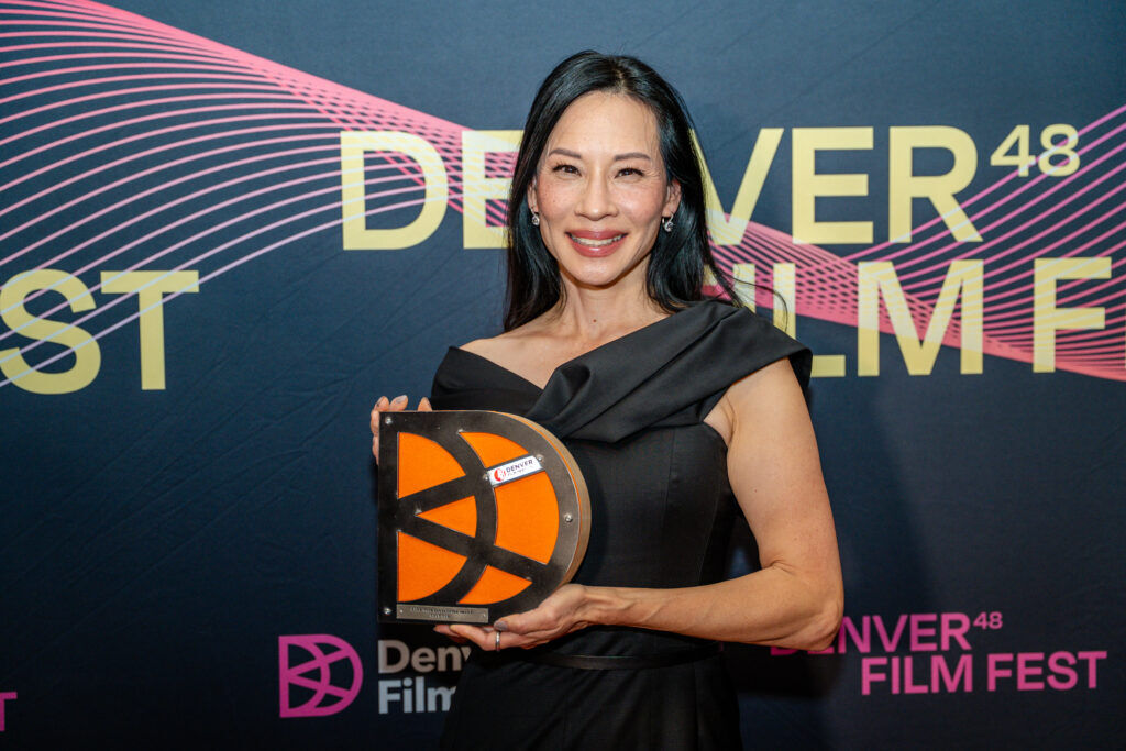 Actor and filmmaker Lucy Liu sits on stage during a Denver Film Festival conversation, smiling while holding a microphone with the festival logo, as her name appears on the screen behind her.