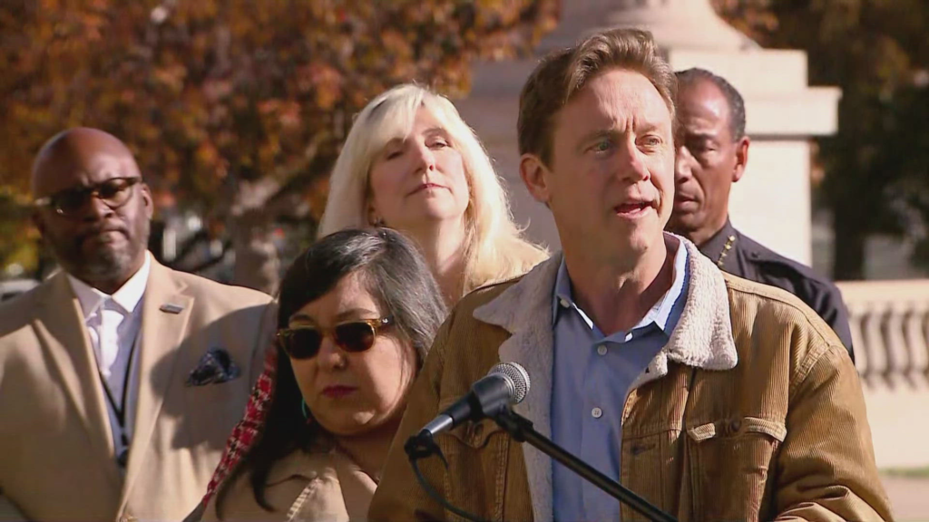 Mayor Mike Johnston speaks at an outdoor press conference with a microphone in front of him. Several people stand behind him, listening attentively, with autumn trees and a public building visible in the background.