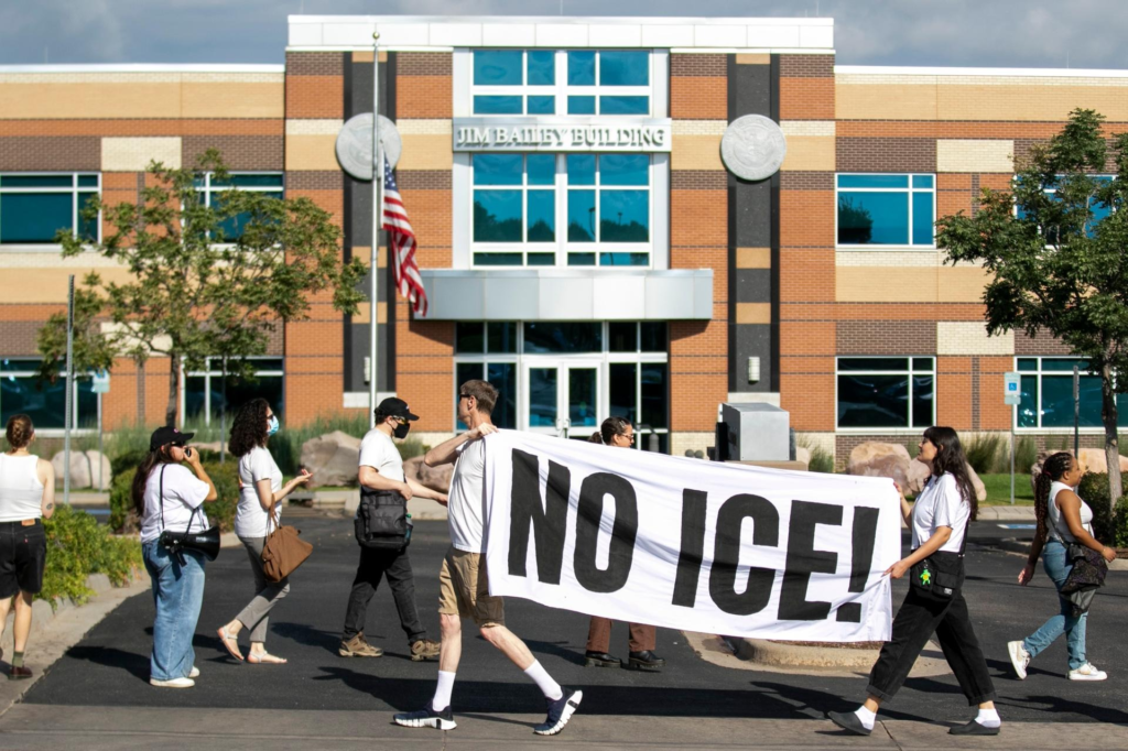 A group of protesters walk past the Jim Bailey Building, holding a large white banner with bold black letters reading “NO ICE!” The U.S. flag flies near the building’s entrance on a sunny day.