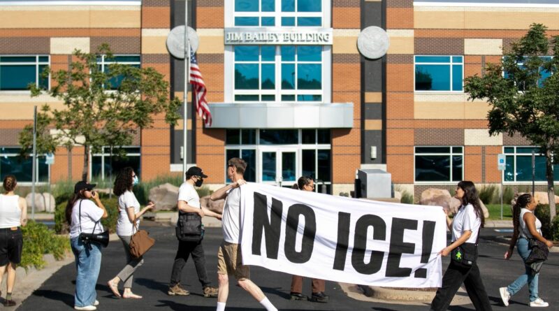 A group of protesters walk past the Jim Bailey Building, holding a large white banner with bold black letters reading “NO ICE!” The U.S. flag flies near the building’s entrance on a sunny day.