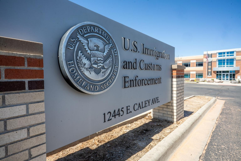 A close-up view of the U.S. Immigration and Customs Enforcement sign outside the Jim Bailey Building, showing the Homeland Security seal and the address “12445 E. Caley Ave.” The brick-and-glass building stands in the background under a clear blue sky.