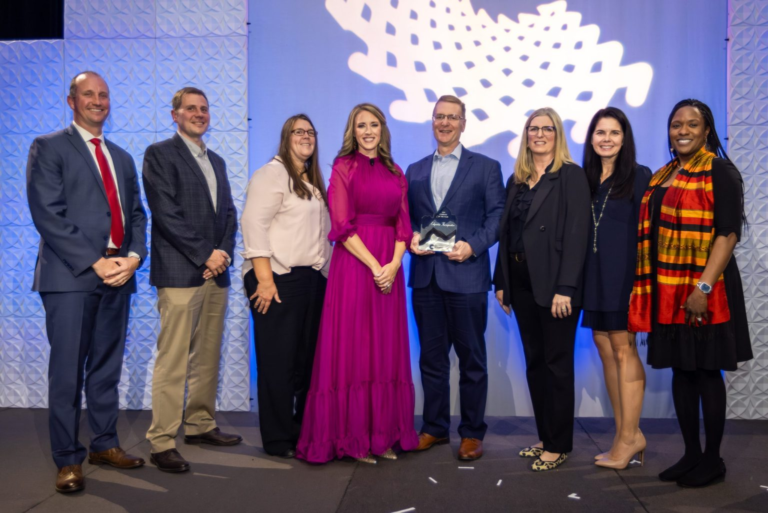 A group of eight people stand together onstage at the Colorado BioScience Association awards event, posing for a photo in front of a geometric white backdrop and blue lighting. One man at the center holds a glass award, while the others smile proudly beside him. The group includes men and women dressed in business and formal attire.
