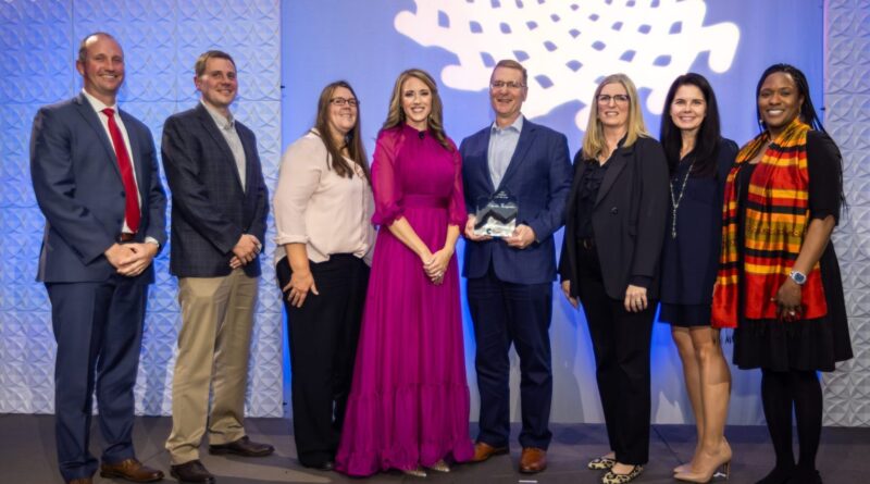 A group of eight people stand together onstage at the Colorado BioScience Association awards event, posing for a photo in front of a geometric white backdrop and blue lighting. One man at the center holds a glass award, while the others smile proudly beside him. The group includes men and women dressed in business and formal attire.