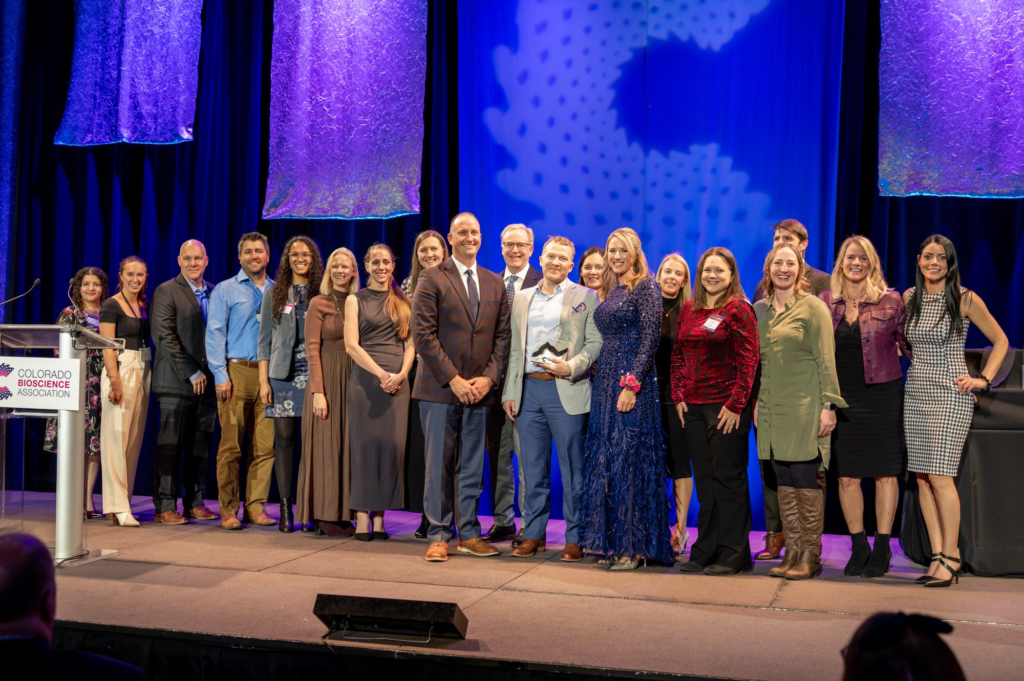 A large group of award recipients and Colorado BioScience Association staff stand together onstage at the 22nd Annual Awards Celebration. They are smiling and posing for a group photo under purple and blue stage lighting, with a podium displaying the Colorado BioScience Association logo on the left.