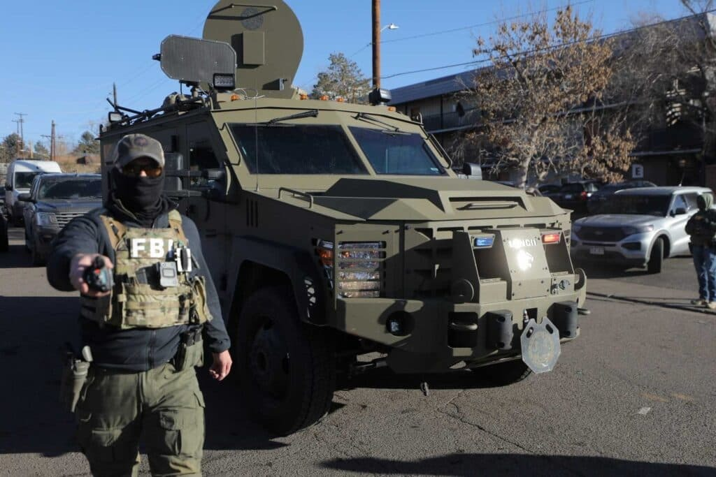 An FBI agent wearing tactical gear and a face covering stands in front of a large armored vehicle parked on a residential street. Several vehicles and a few bystanders are visible behind him on a cold, sunny day.