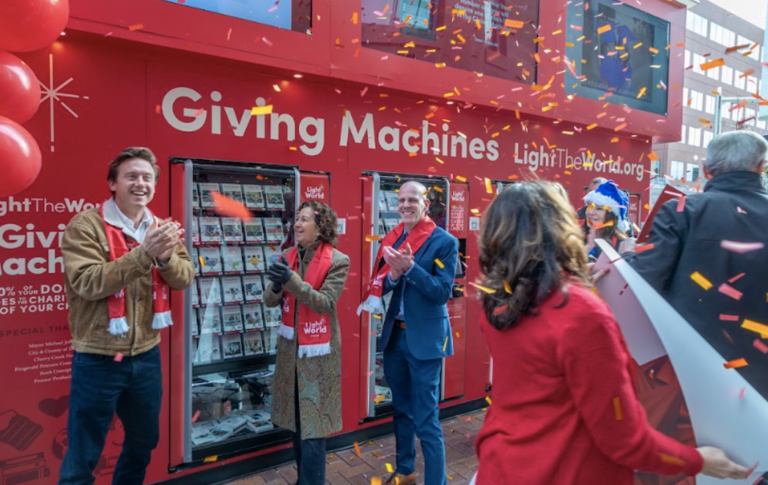 A festive outdoor celebration at the Light the World Giving Machines in Denver. Mayor Mike Johnston and several community members stand in front of bright red Giving Machines, smiling and clapping as colorful confetti falls around them. All are bundled in winter clothing and wearing red Light the World scarves. Large red balloons and holiday decorations frame the scene.