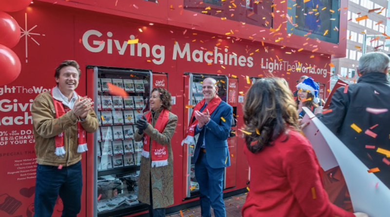 A festive outdoor celebration at the Light the World Giving Machines in Denver. Mayor Mike Johnston and several community members stand in front of bright red Giving Machines, smiling and clapping as colorful confetti falls around them. All are bundled in winter clothing and wearing red Light the World scarves. Large red balloons and holiday decorations frame the scene.