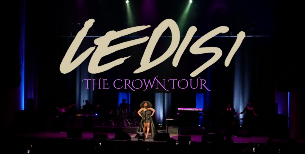 Ledisi performs center stage at the Paramount Theatre, standing under colorful overhead lights as audience members record with their phones. The stage is framed by blue and purple lighting, with the band positioned behind her.