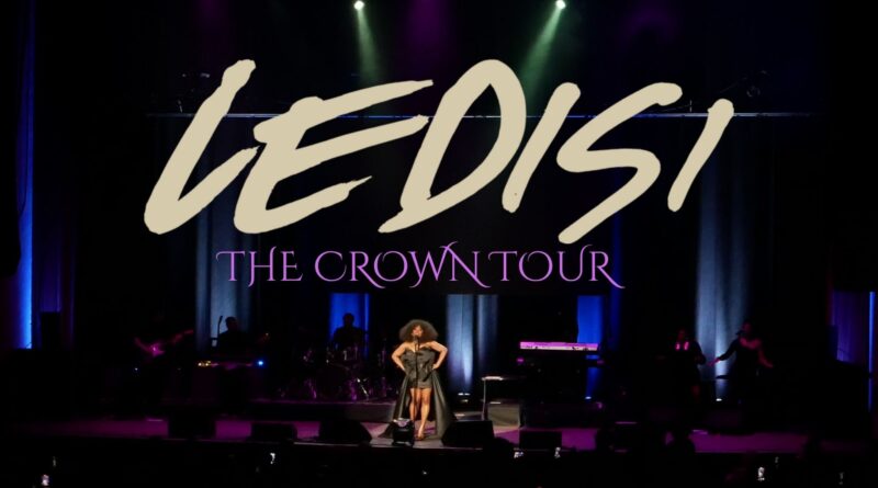 Ledisi performs center stage at the Paramount Theatre, standing under colorful overhead lights as audience members record with their phones. The stage is framed by blue and purple lighting, with the band positioned behind her.