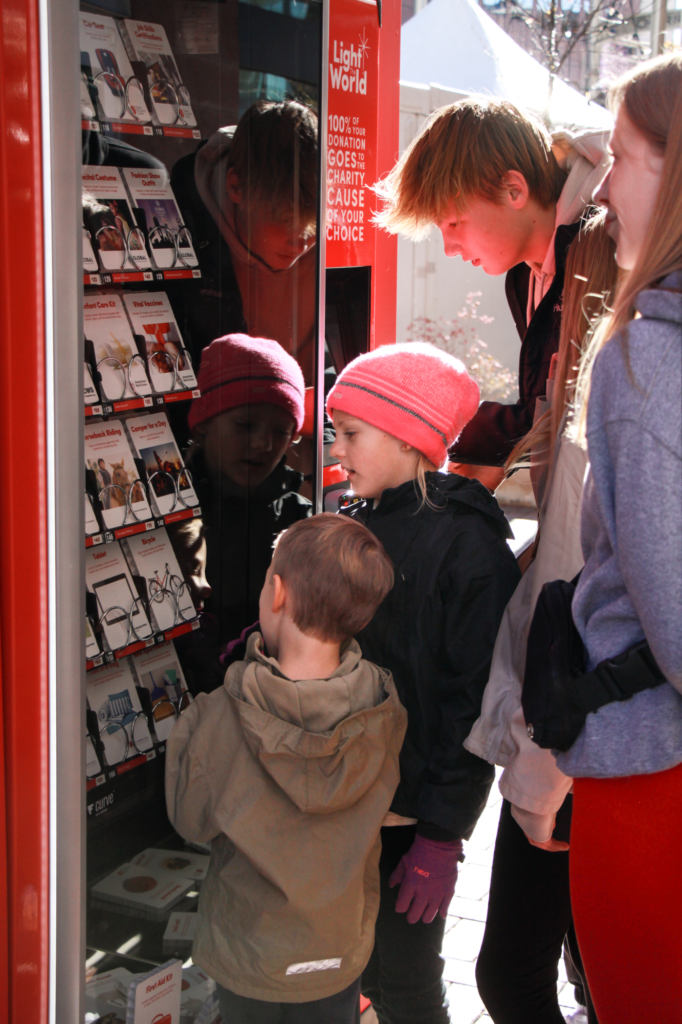 A group of children huddles around a Giving Machine, intently examining the donation item cards displayed inside. A young girl in a bright pink beanie points toward one of the selections while a smaller child in a brown jacket looks closely at the options. Sunlight reflects on the machine’s glass as the kids explore items like hygiene kits, medical supplies, and food donations.
