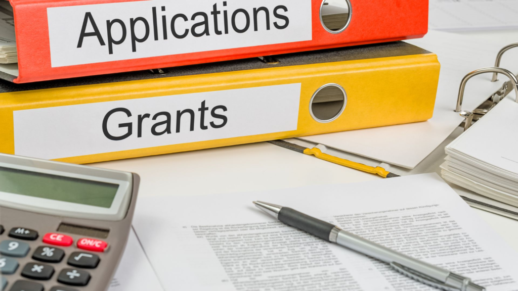 Two large binders labeled “Applications” and “Grants” sit stacked on a desk surrounded by paperwork, a calculator, and a pen, representing the administrative process of applying for nonprofit or government funding.
