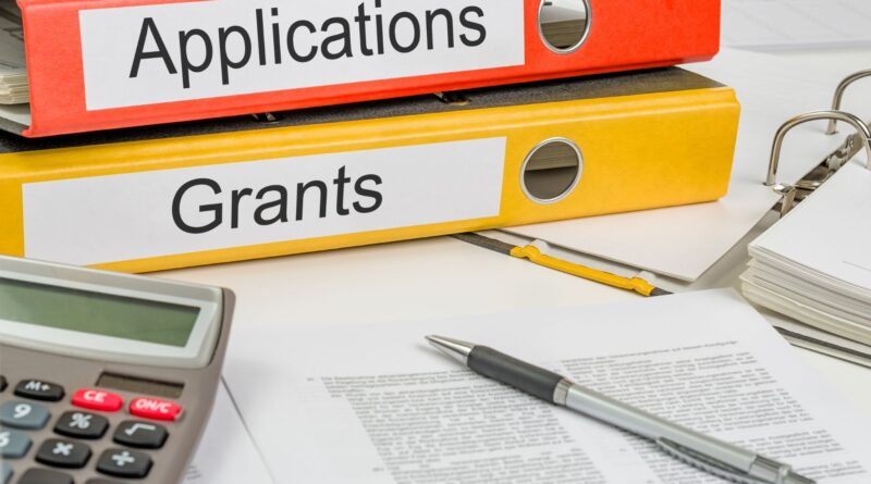 Two large binders labeled “Applications” and “Grants” sit stacked on a desk surrounded by paperwork, a calculator, and a pen, representing the administrative process of applying for nonprofit or government funding.