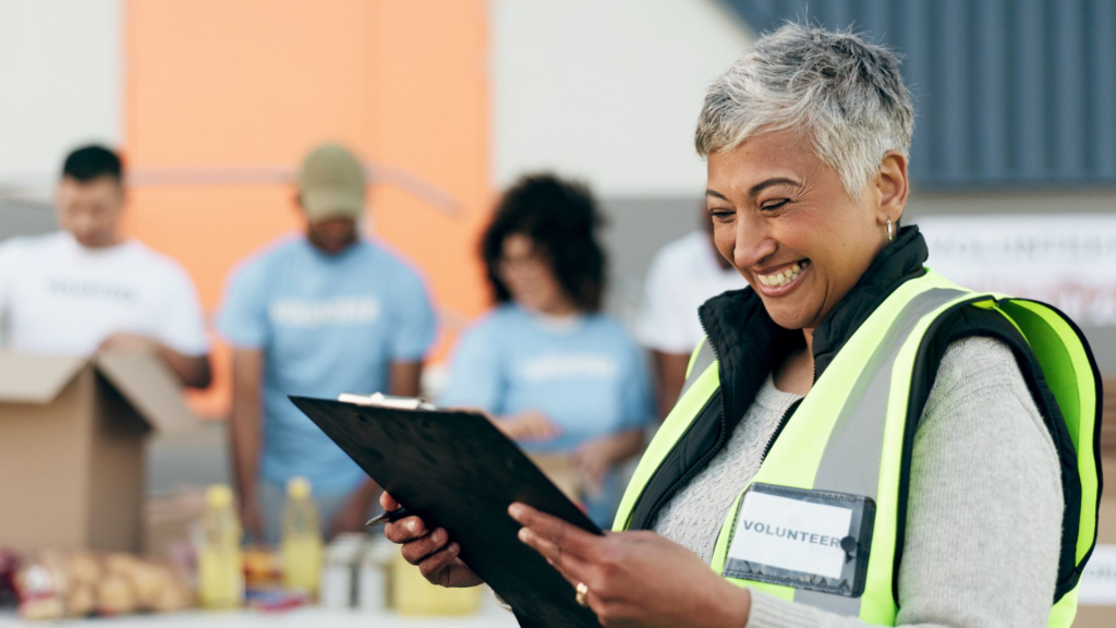 A smiling woman with short gray hair wearing a yellow safety vest and holding a clipboard stands outdoors at a volunteer event. Behind her, several volunteers in light blue shirts sort food and supplies on a table.