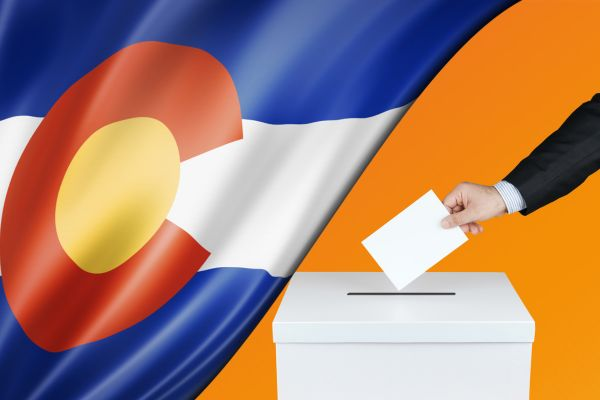 A person casts a ballot into a white voting box against a backdrop featuring the Colorado state flag, symbolizing civic participation and elections in Colorado.