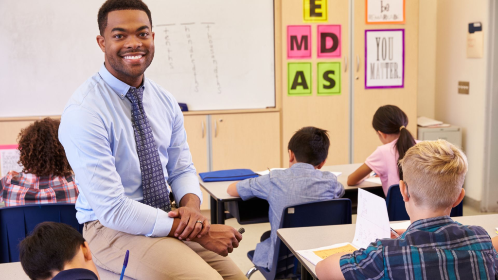 A smiling teacher sits on a desk in an elementary school classroom while students work at their desks. The teacher, dressed in a light blue shirt and tie, faces the camera as colorful educational posters decorate the wall behind him.