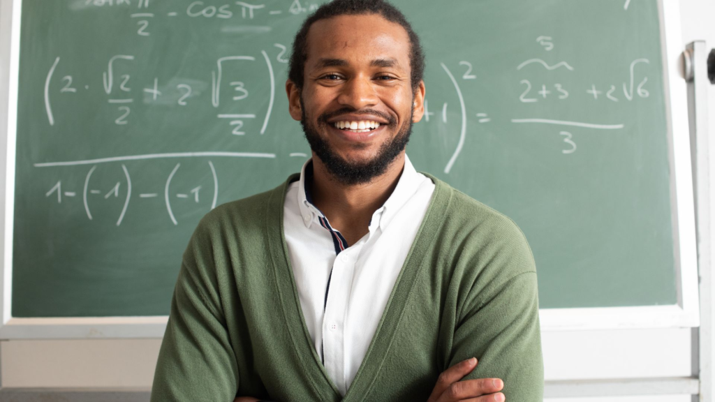 A smiling man stands in front of a chalkboard filled with math equations. He wears a green sweater over a white collared shirt, with his arms folded confidently across his chest, appearing as a teacher or professor in a classroom setting.