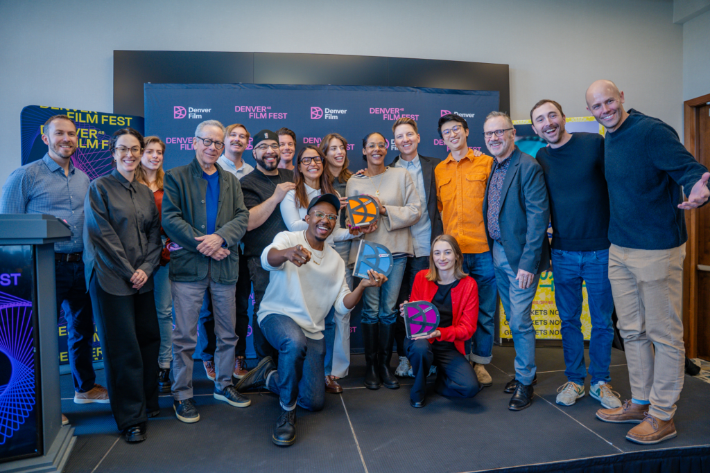 A group of filmmakers, jurors, and award recipients pose together during the Denver Film Festival awards brunch, smiling and holding colorful DFF48 trophies in front of a branded backdrop.