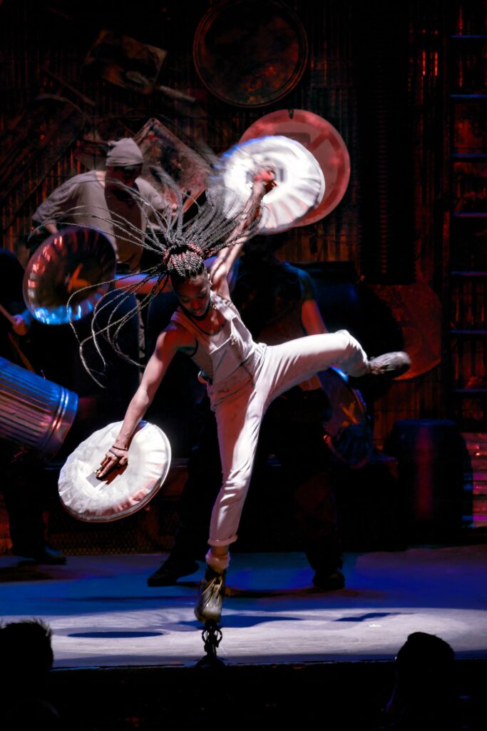 A performer in white overalls leaps midair while playing metal trash can lids, braids flying dramatically as other cast members drum behind her on a dimly lit industrial set.