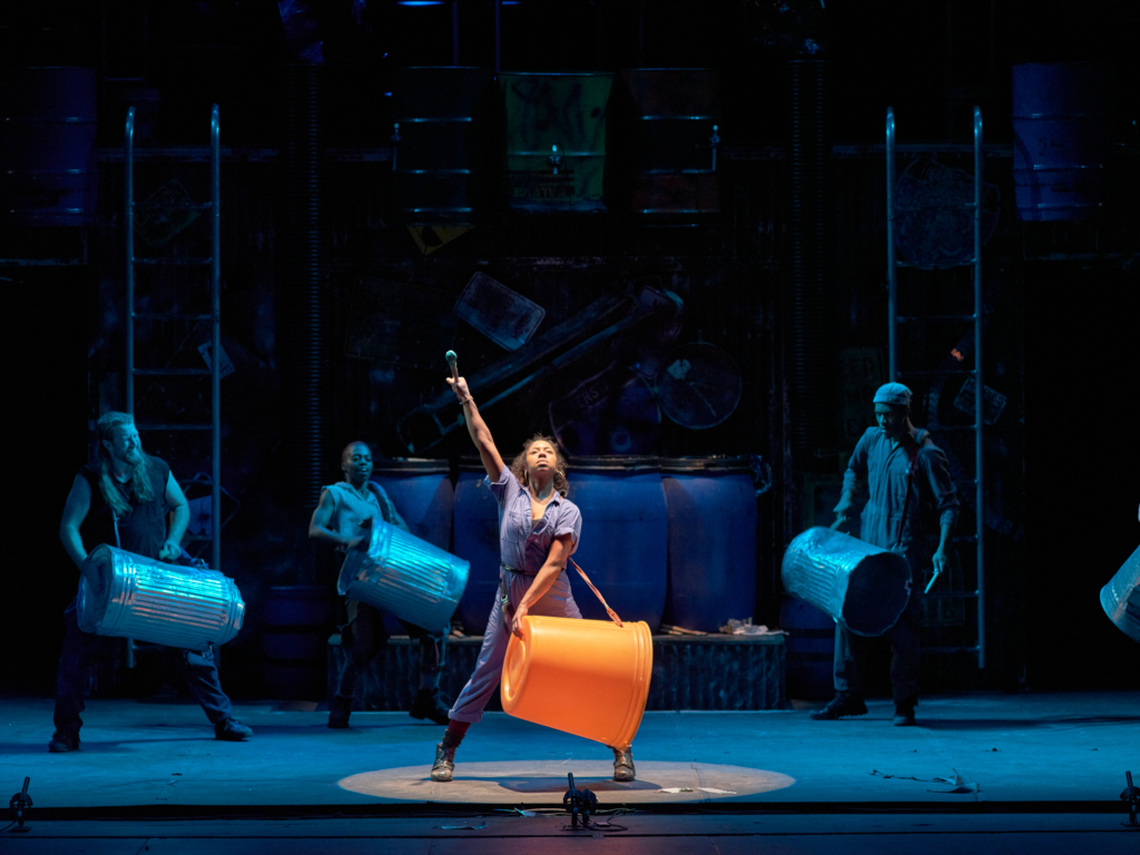 A performer in a blue jumpsuit raises one arm triumphantly while holding an orange plastic bin, surrounded by cast members rhythmically striking metal trash cans under blue-toned lighting.