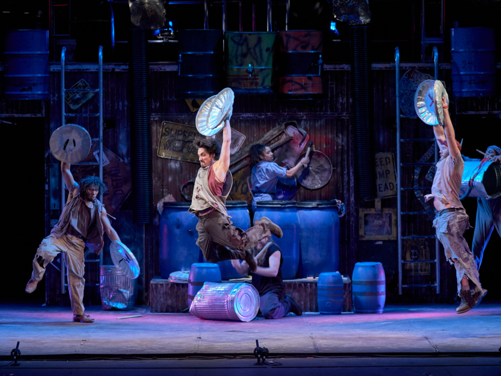 Three performers leap with metal trash can lids raised overhead while others drum on large barrels in front of a towering backdrop made of rusted metal, pipes, and industrial debris.