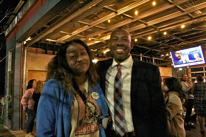 Rob Andrews and Alli Jackson smile and pose for a photo at an evening election-night gathering, standing under string lights with other attendees and a television screen in the background.