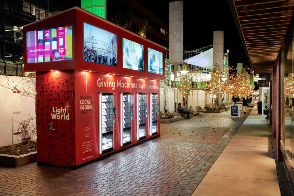 A row of illuminated Light the World Giving Machines glows along a brick walkway at night in Cherry Creek North. Twinkling holiday lights hang overhead and wrap around nearby trees, creating a festive atmosphere. Digital screens on top of the red machines display animated visuals, while a few evening visitors walk through the decorated plaza.
