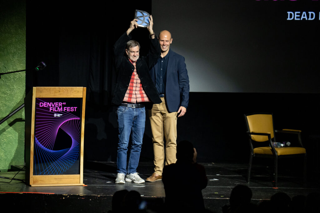 Film director Gus Van Sant stands on stage at the 48th Denver Film Festival holding his award aloft beside a presenter, as the audience applauds his recognition for Excellence in Directing.