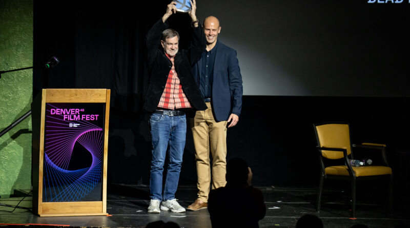 Film director Gus Van Sant stands on stage at the 48th Denver Film Festival holding his award aloft beside a presenter, as the audience applauds his recognition for Excellence in Directing.