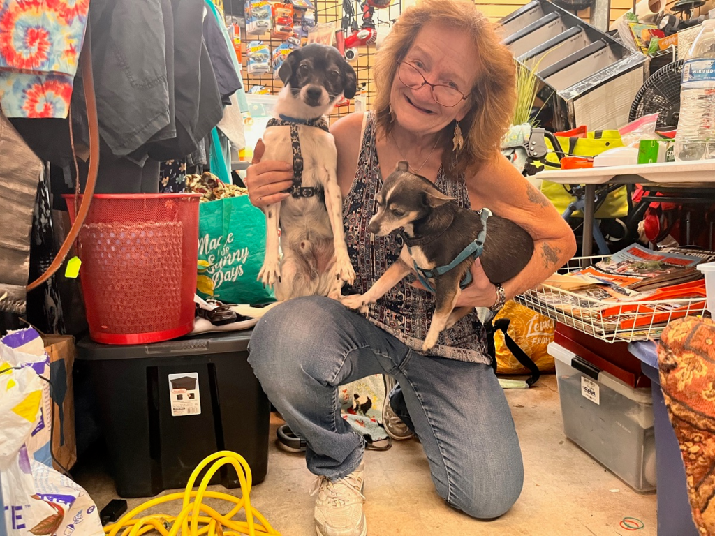 A smiling woman kneels inside a cluttered indoor space, holding two small dogs in harnesses—one in her arms and the other standing upright on its hind legs. She is surrounded by bags, boxes, clothing, and miscellaneous household items.