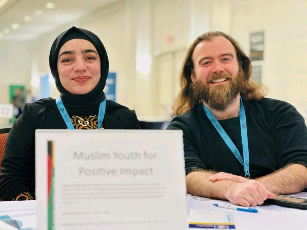 Two people sit at a table with a sign reading “Muslim Youth for Positive Impact.” One is a woman wearing a black hijab and smiling warmly, and the other is a man with long hair and a beard who is also smiling. Both wear blue conference lanyards.